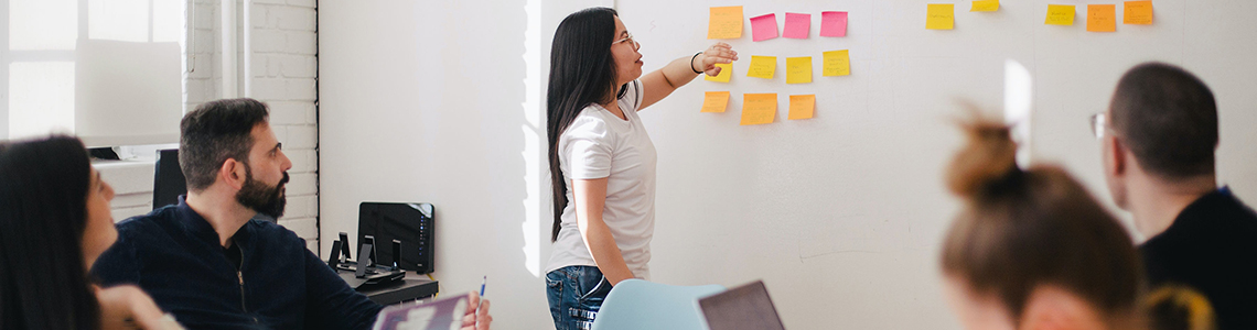 Photo of a small group of people sat around a table with a person stood up putting a post-note on a whiteboard.